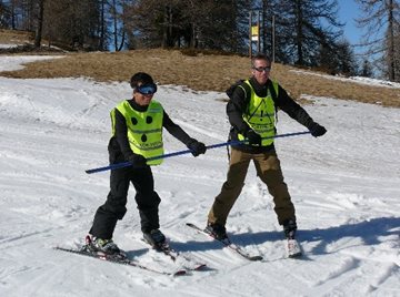 Voorbeeld van begleider en vb-skier die naast elkaar skien en samen
gebruik maken van één slalomstang. Zij houden de slalomstang allebei voor de
borst met twee handen vast.