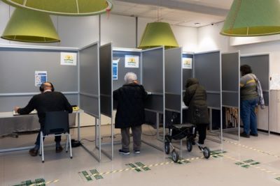 Stemlokaal met vier stemhokjes. In het linker stemhokje is een man met stemmal
en stembox aan het stemmen.